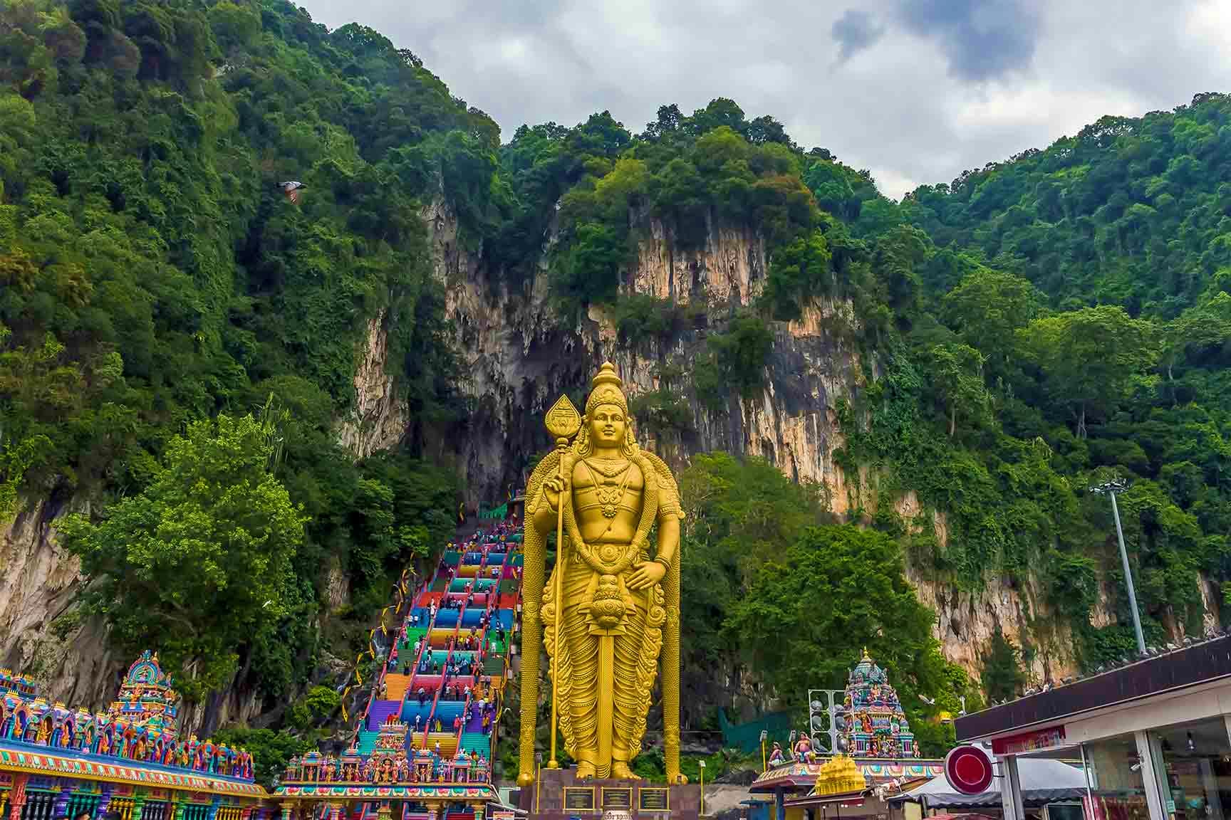 Large golden Hindu deity statue at Batu Caves entrance with colorful staircase and limestone cliff in Malaysia